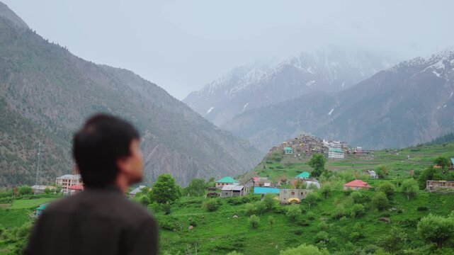 Defocused Indian man looking at Triloknath village in front of the snowy Himalayan mountain peaks at Pattan valley in Lahaul, Himachal Pradesh, India. Man looking at the village on cliff during summer