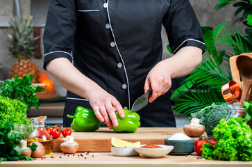 Chef cuts green bell pepper. Cozy kitchen with wooden table, kitchenware, vegetables, herbs and ingredients for cooking. Healthy vegan eating, culinary, recipes, food blogging