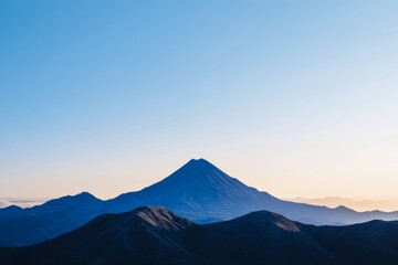 Majestic volcano rises above rolling hills at dawn under a clear blue sky with a hint of orange