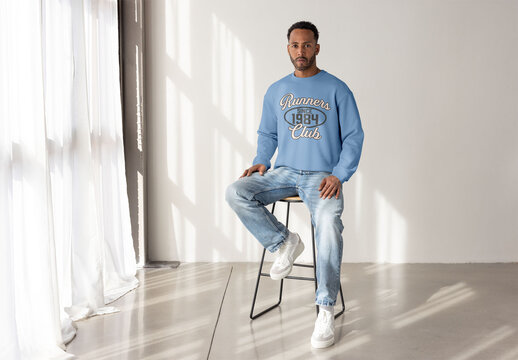 Mockup of black man in a blue sweater looks confidently at the camera while sitting on a stool in a bright studio setting.