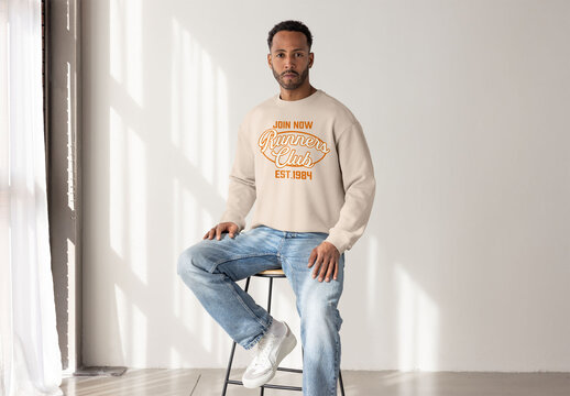 Mockup ofBlack man wearing a sweater sits on a stool in a bright studio, looking directly at the camera 