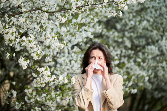 Woman allergic suffering from seasonal allergy at spring in blossoming garden at springtime. Young woman sneezing and blowing nose with nasal handkerchief in front of blooming tree. Allergy concept.