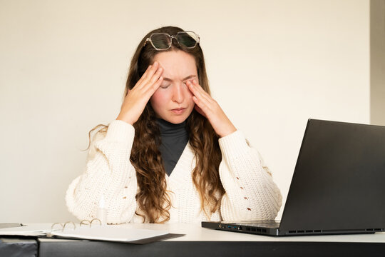 A young tired woman with glasses working on a laptop having a painful headache touching her temples. Female student with stress. Migraine, stress relief, chronic pain, digital eye strain symptoms.