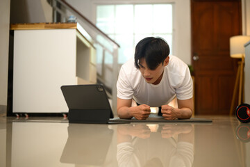 Focused young man doing plank exercise on a mat and watching training videos on digital tablet