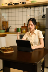 Happy woman sitting at the dining table sipping coffee and using digital tablet