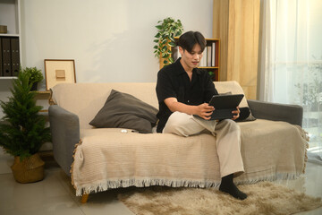 Obraz premium Relaxed asian man sitting on cozy sofa in a modern living room and using digital tablet
