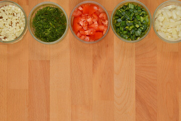 Five glass bowls containing chopped vegetables - onions, leeks, tomatoes, dill, and garlic - are...