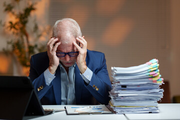 Senior businessman having a headache from excessive workload, sitting at his desk with hands on his...
