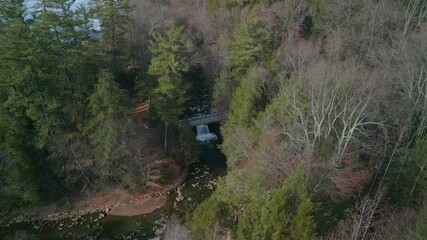 Aerial drone view of a waterfall and bridge in between trees.