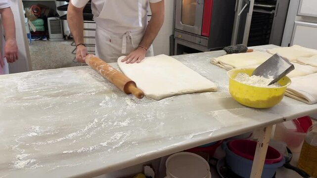 Young caucasian pastry chef rolling puff pastry dough on a white mable table inside a traditional spanish bakery. 4K