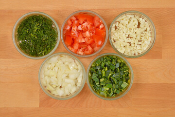 Five glass bowls containing chopped vegetables - dill, tomatoes, garlic, onions, and leeks - are...