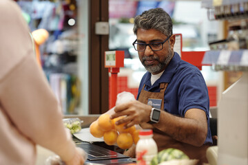 Portrait of Indian man assisting customer and weighing bag of oranges while working as cashier at...