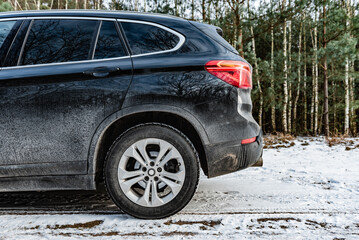 Very dirty salty black car is parked on nature covered by snow in winter.