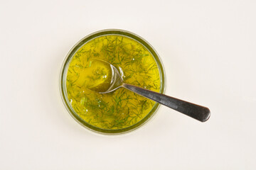 A top-down view of a clear glass bowl filled with olive oil, dill, and lemon juice, with a silver spoon resting inside