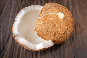 A cracked coconut shell on a rustic wooden table. A key ingredient in Indonesian cuisine, often used in Eid dishes like opor ayam. The rough brown husk contrasts with the smooth white flesh inside