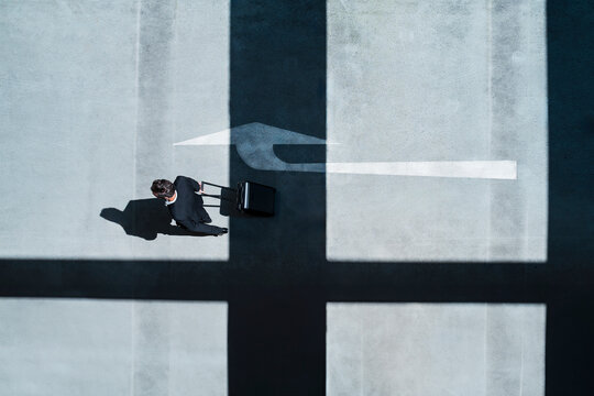 Businessman in suit with suitcase walking on concrete surface with arrow shadow
