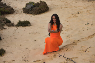 Young, pretty brunette woman in orange dress, bending down, holding a handful of sand and falling...