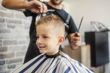 Head shot of a smiling little boy sitting in barber shop while hairdresser cutting his hair with...