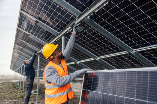 Interracial worker with drill in hand installing solar panels at solar farm.