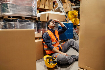 Exhausted interracial warehouseman sitting on floor surrounded by boxes and rubbing his painful...