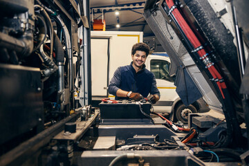 Portrait of happy diverse mechanic using battery charger on truck at mechanic workshop.