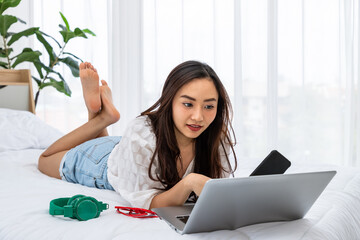 Asian woman working on laptop while relaxing on bed in cozy bedroom