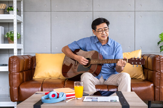 Senior Asian man playing acoustic guitar - relaxing on leather sofa with music and coffee table essentials