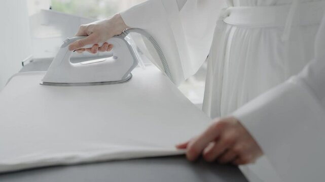 A person dressed in white using a steam iron to press a white garment on an ironing board. The setting suggests domestic chores and household routine. Person Ironing White Garment at Home
