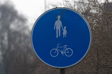 A circular blue sign indicates shared paths for pedestrians and cyclists, featuring icons of a man and child walking, alongside a bicycle symbol. The environment is quiet, suggesting early morning