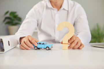 Cropped shot of a car insurance agent sitting at an office desk and holding a little blue toy automobile model and a wooden question mark sign in his hands. Car insurance business concept