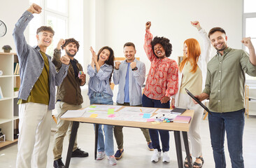 Cheerful group of diverse company employees standing together next to desk and raising hands. Multiracial team of colleagues celebrating successful teamwork, happy from excellent results of work.