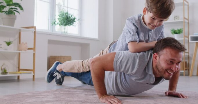 Athletic cheerful father exercising at home, doing push ups in living room with little boy on his back. Strong dad and his smiling son having fun, playing sports together at home. Family workout.