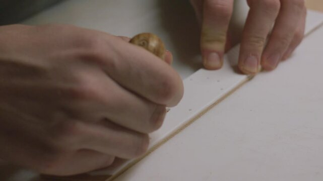 A detailed close-up of a craftsman carefully marking leather with an awl using a template for precision.