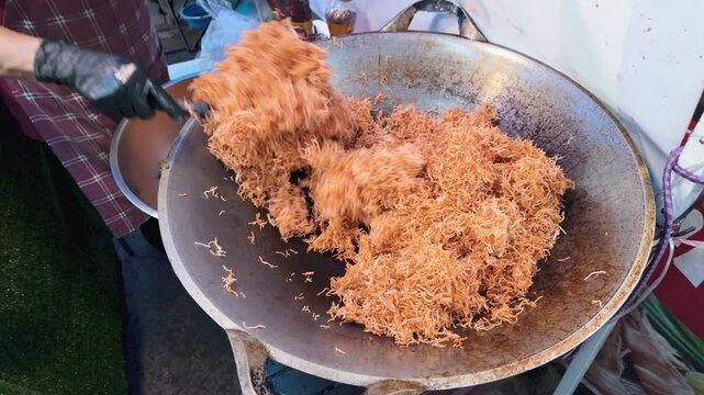 Crispy Thai Kanom Mee Krob rice noodles being deep-fried in a large wok at a street food stall, then scooped and transferred into a stainless steel mixing bowl for further preparation View from above