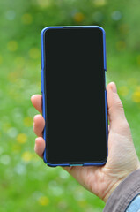 Hand with smartphone mockup against fresh green background. Phone with black screen template in hand .Closeup outdoors photo. Free copy space.