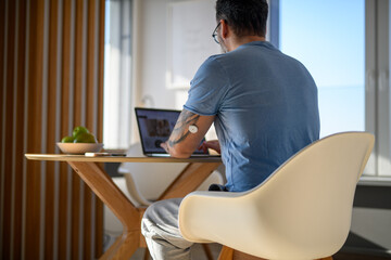 Man with CGM sensor on arm sitting at table and working on laptop.