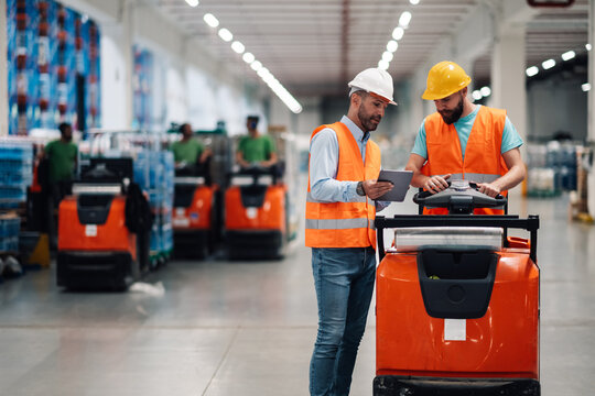 Warehouse workers using tablet and driving tow tractor with merchandise