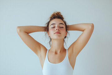 Fototapeta premium A girl wearing a white tank top stretches her arms in a relaxed pose against a white studio background, exuding a sense of comfort and ease.