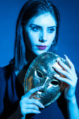 Naklejka premium The girl is holding a golden mask in her hands. Studio photo of a girl with blue light.