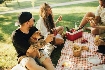 Happy people having picnic with hot dogs and cold drinks, sitting on a plaid in a park.