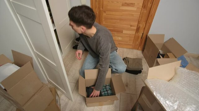 Young man in living room and packing clothing into cardboard box before moving. Surrounding him are multiple boxes, scattered clothes, and packing materials in a cluttered room.