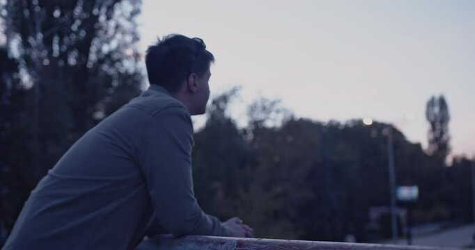 Contemplative Young Man Leans on Bridge Railing, Watching City at Dusk