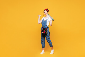 Fototapeta premium Full body young employee laborer handyman woman wears overalls belt hold spirit level show thumb up isolated on plain yellow background. Instruments for renovation apartment room. Repair home concept.