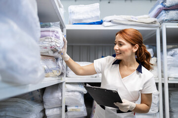 Female laundry worker standing in dry cleaning service storage room. In background, clean folded...