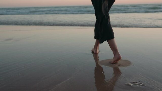 Woman in a black dress walks along a beach at sunset, waves gently lapping at the shore. Perfect for travel, vacation, or mindfulness videos.