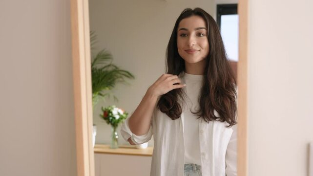 A beautiful brunette young woman fixes her hair while looking into a mirror, preparing to go out.