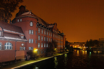 Fototapeta premium Orange hues envelop the historic Saint Thomas school building along the Ill River, viewed from Pont Saint-Martin at night.