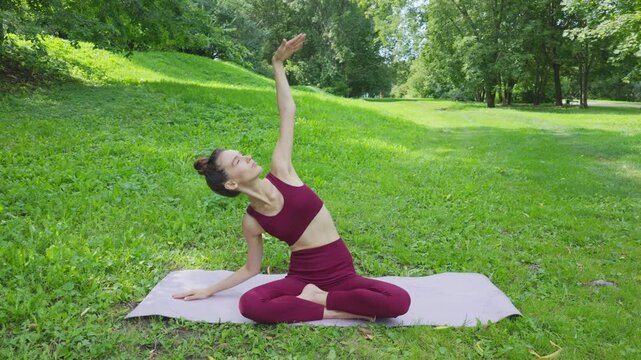 Beautiful young fit brunette doing stretching and yoga on the mat in the open aii in the green park in summer