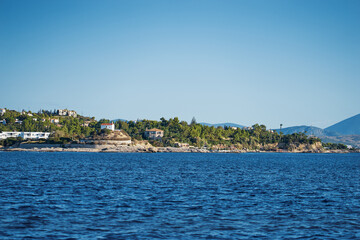 Naklejka premium A beautiful coastal view of Greece island showcasing vibrant blue waters, lush greenery, and idyllic residential areas under a clear blue sky, conveying serenity and natural beauty.