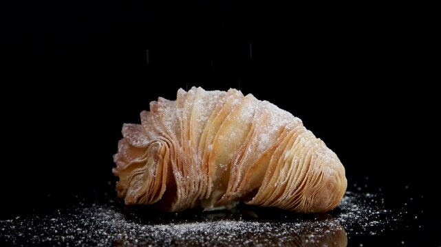 A 360-degree rotating view of a Sfogliatine alla Crema croissant on a black background, with powdered sugar delicately falling on the flaky pastry, highlighting its layers and creamy filling.
